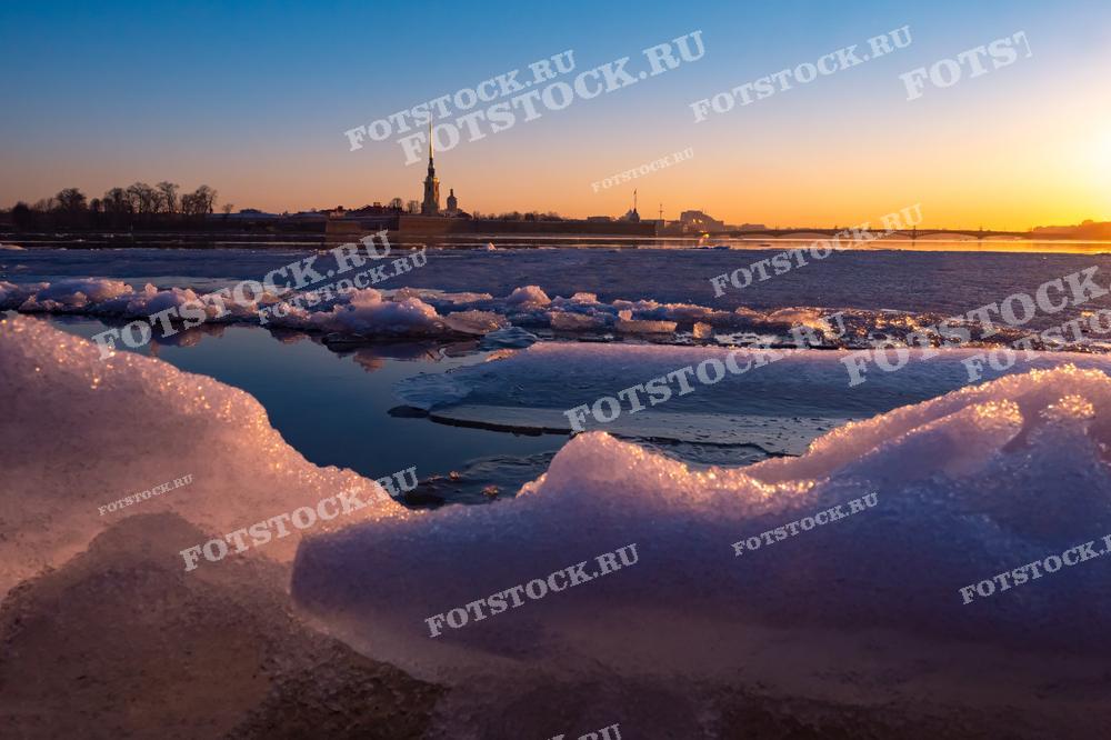 На фотографии представлена весенняя панорама на Неву в Санкт-Петербурге. Тающий снег и лед окрашенны в тёплые оттенки заходящего солнца. На заднем плане виднеется силуэт Петропавловской крепости, что придаёт кадру историческую значимость. Эта фотография может быть использована в публикациях о зимнем отдыхе в России, для статей о природе и климате страны или как часть художественной экспозиции, демонстрирующей красоту российских городов.